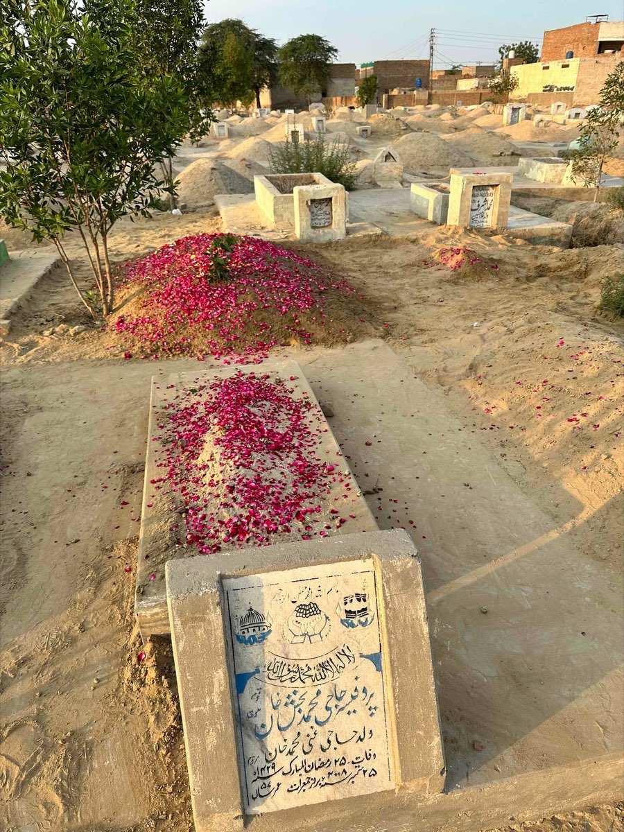 Two graves side by side in a Pakistani cemetery at dusk, the larger covered with red rose petals on a sand mound, the smaller a flat stone slab with rose petals scattered across, an Urdu-inscribed headstone in the foreground, a small green tree to the left, other plain sand graves visible in the background.