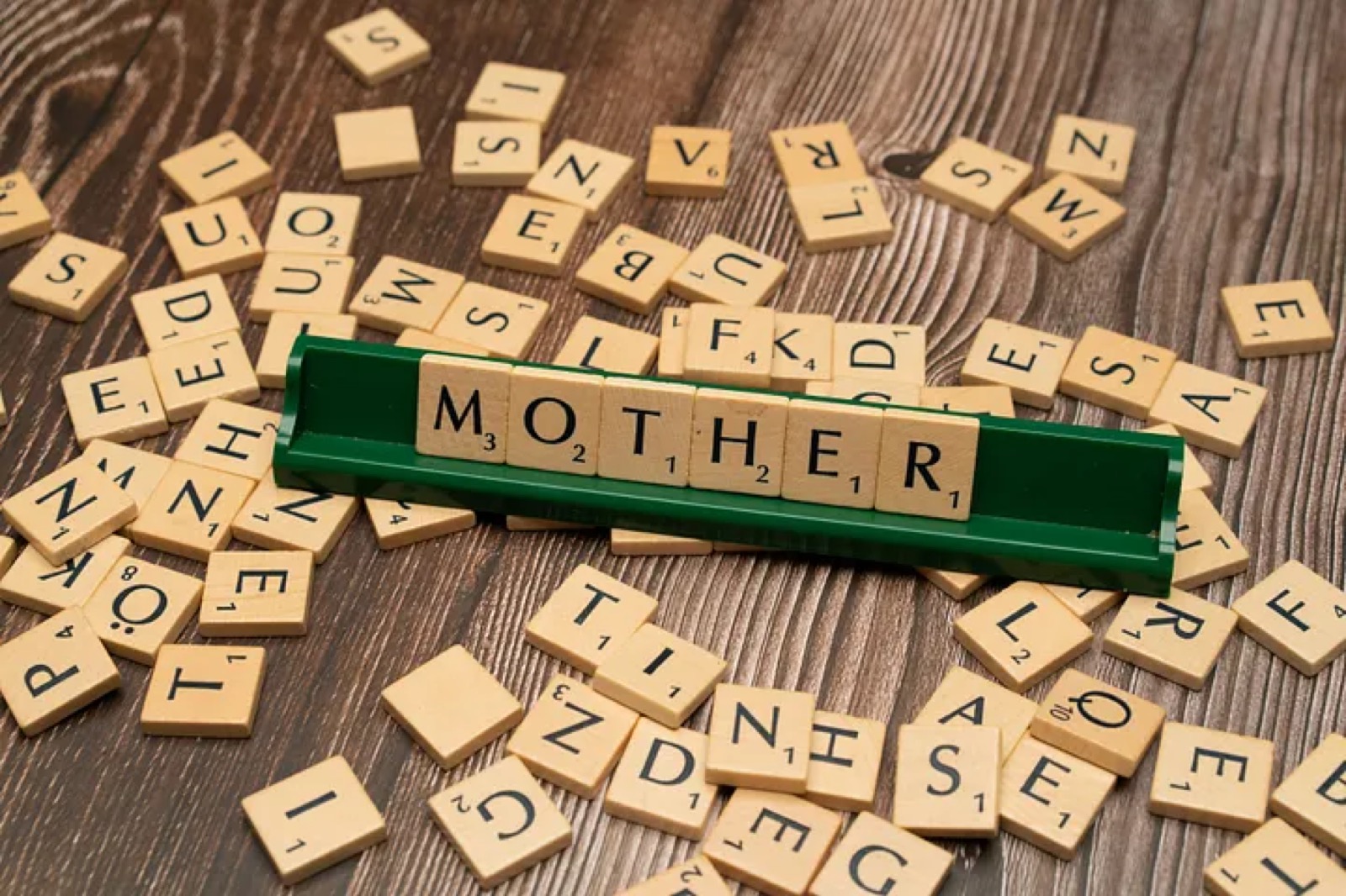 Wooden Scrabble tiles scattered on a dark wood surface, with green tile holders at the centre spelling out the word MOTHER in larger raised tiles.